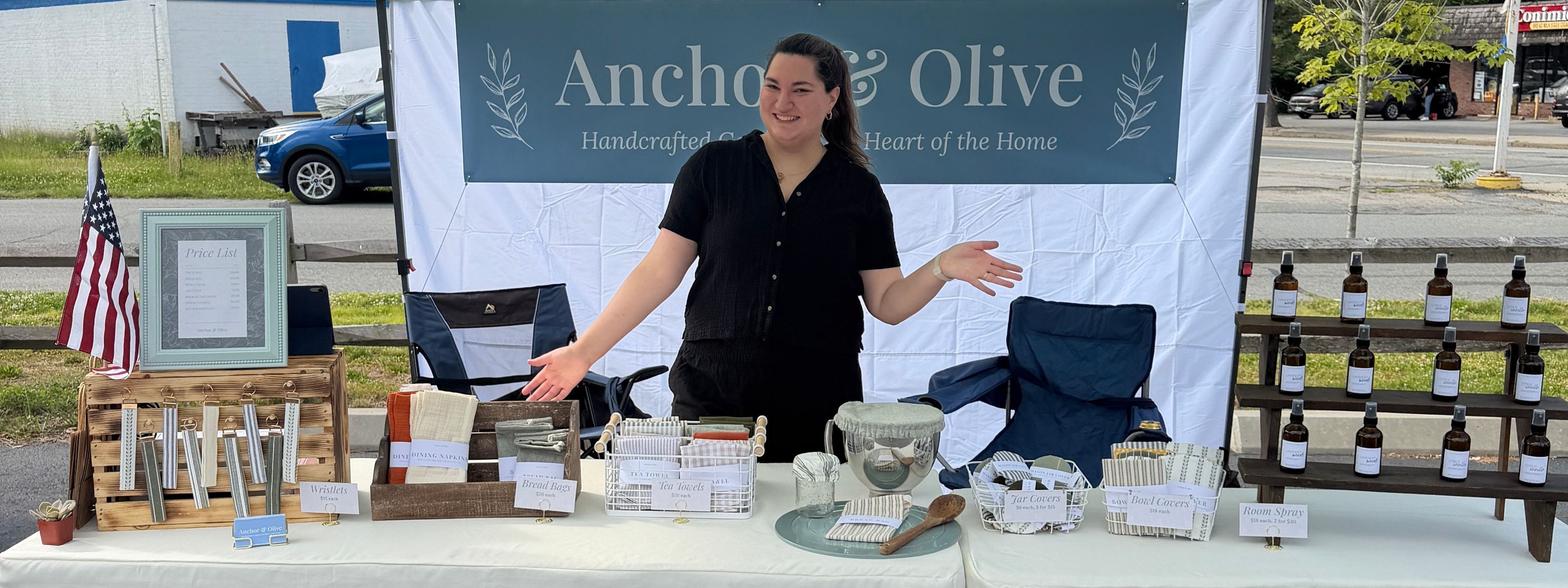Person standing behind a table with various items at an outdoor event under a tent with 'Anchor & Olive' branding.